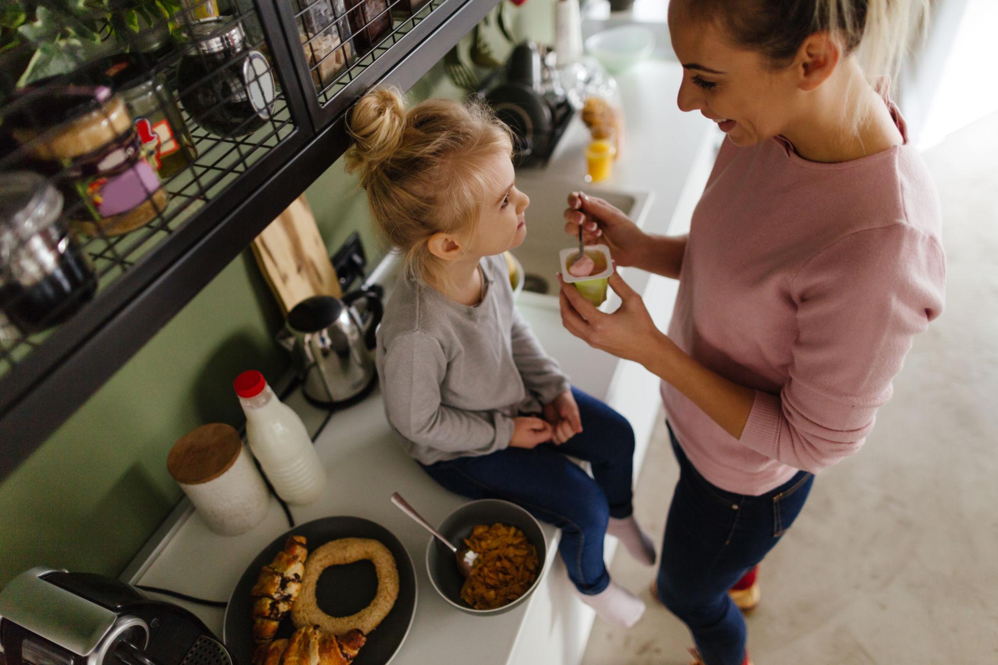 Easy Vitamin D3 for Kids at Breakfast Woman gives a young child a spoonful of yogurt at the kitchen counter during breakfast.